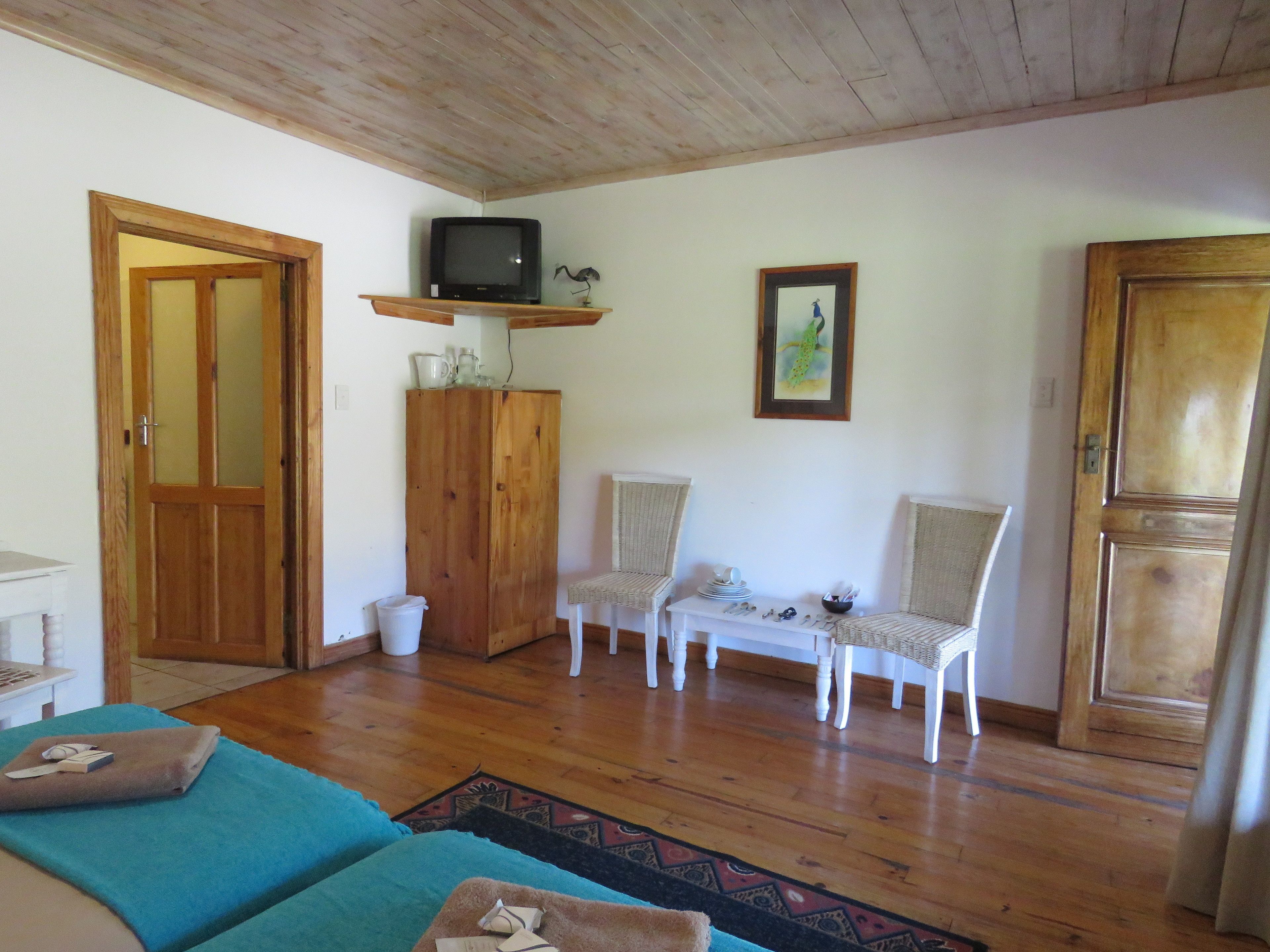 A room with wooden flooring, a television on a shelf, two wicker chairs, a small table with dishes, and a door with a mirror.