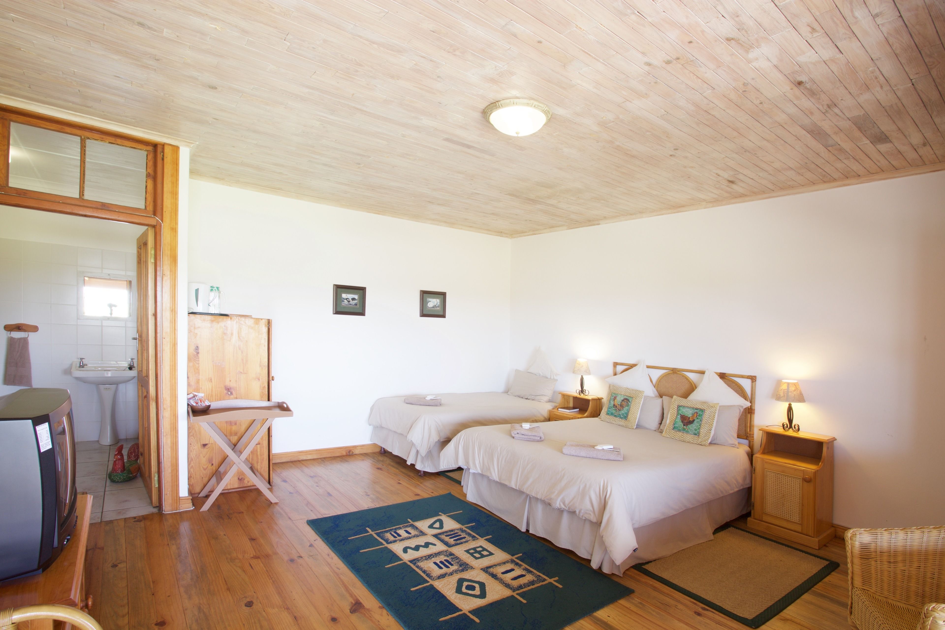 A bedroom with two beds, a wooden ceiling, a small table, a chair, and a bathroom visible through an open door.