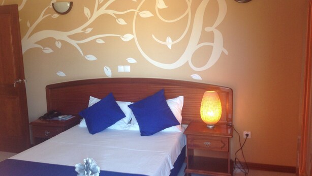 Un lit bien fait, avec des oreillers bleus et un couvre-lit blanc, une lampe de chevet et un mur orné d’un grand motif en forme de lettre « B ».