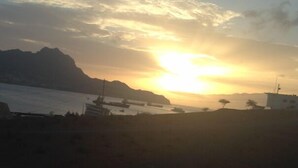 Un coucher de soleil sur une zone côtière, avec des montagnes en arrière-plan et quelques bâtiments près du rivage.