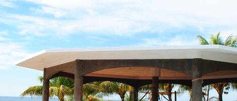 Un área de estar al aire libre techada con piscina, palmeras y vista al océano.