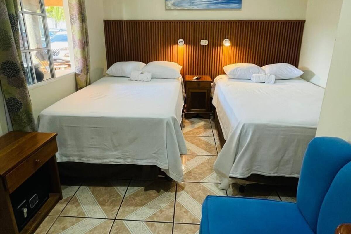 Hotelkamer met twee bedden, een blauwe stoel, een houten nachtkastje, een schilderij aan de muur en een raam met gordijnen.