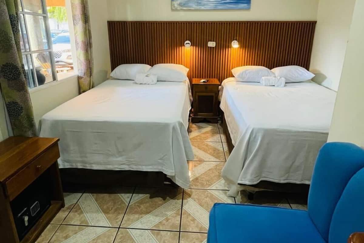 Hotellihuone, jossa on kaksi sänkyä, sininen tuoli, puinen yöpöytä, seinällä oleva taulu ja ikkuna, jossa on verhot.