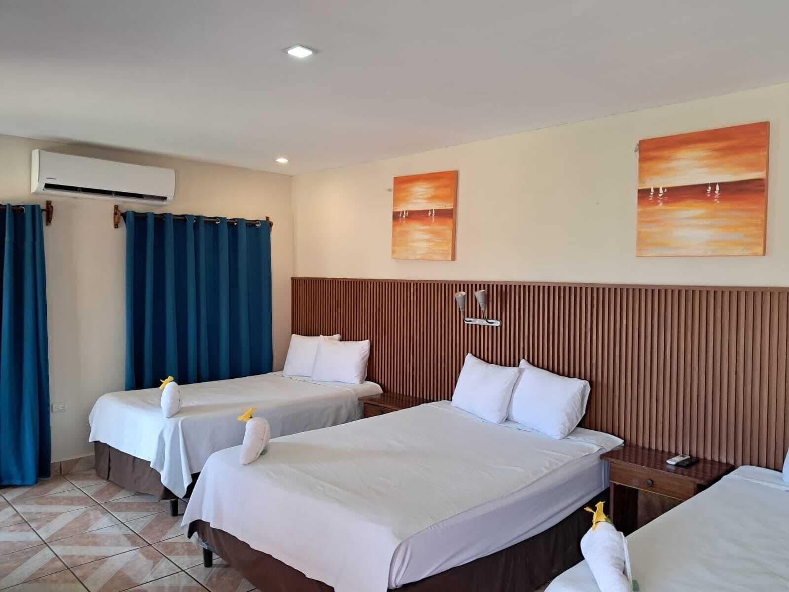 Habitación de hotel con dos camas, cabecero de madera, aire acondicionado de pared y dos cuadros.