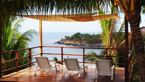 A wooden deck with three white chairs overlooking a beach with palm trees and a rocky island in the distance.