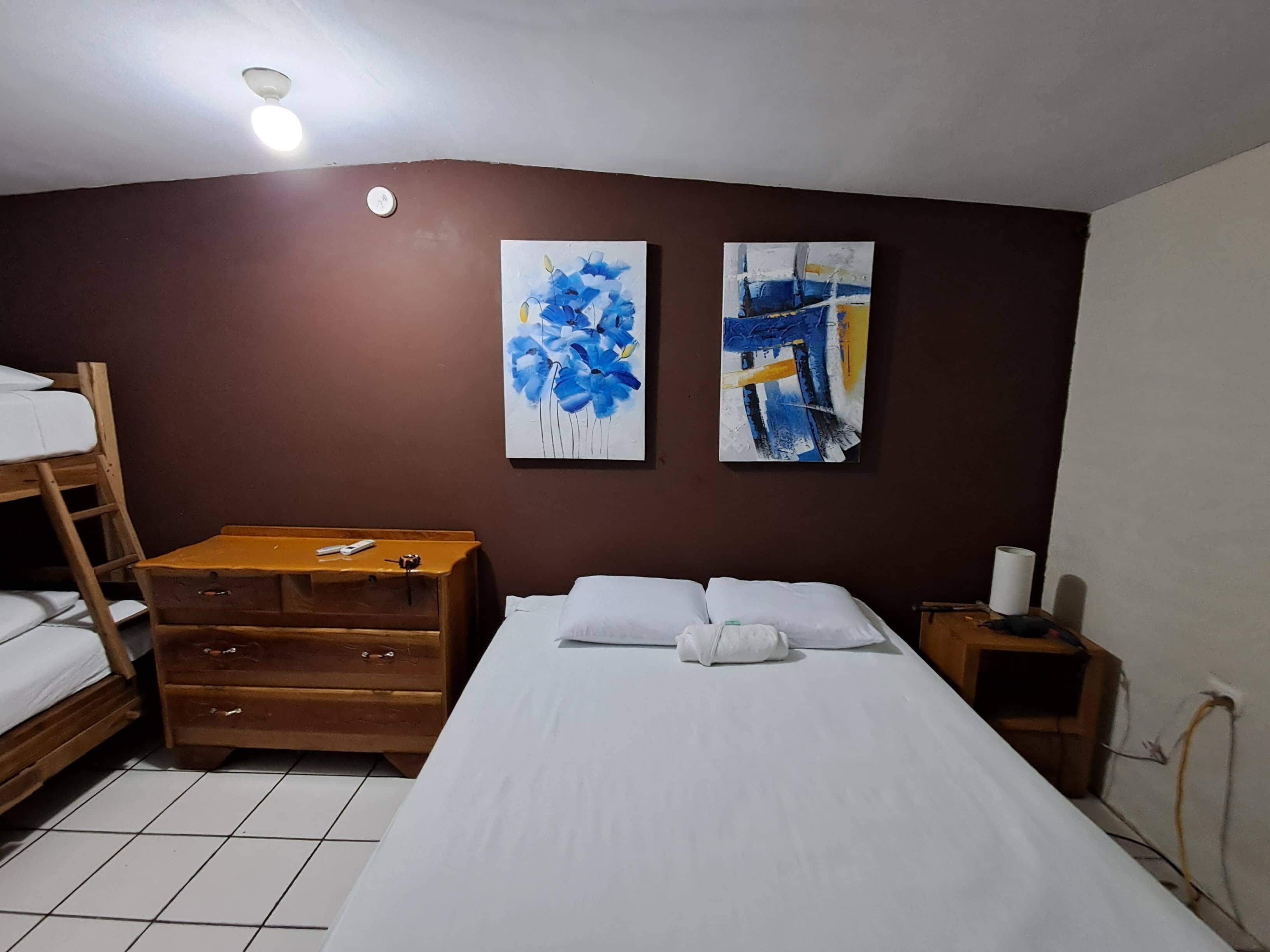 Una habitación con una cama litera, un aparador de madera, un escritorio, dos cuadros abstractos y una lámpara en el techo.