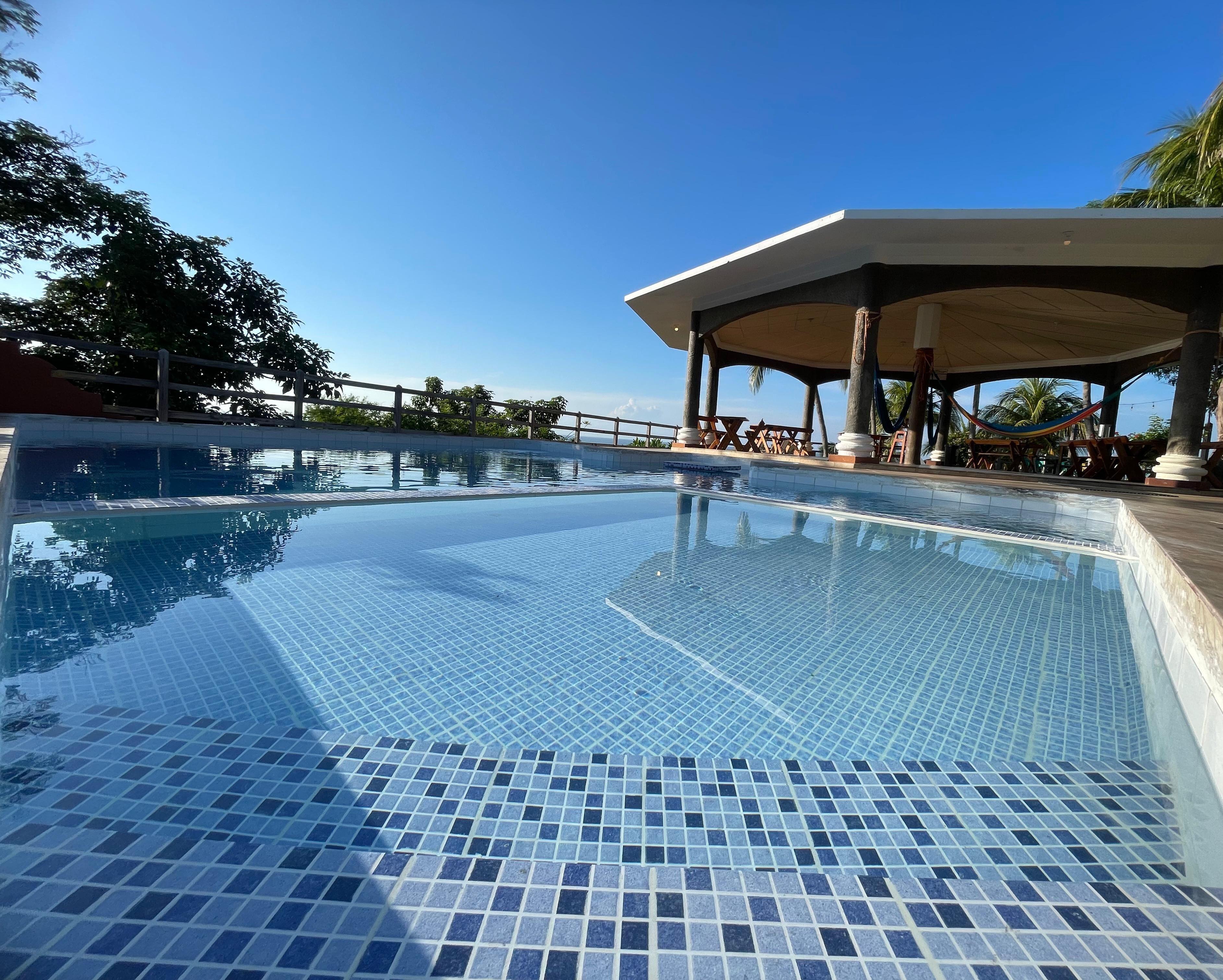 A clear swimming pool with tiled flooring, a covered seating area, and palm trees in the background.