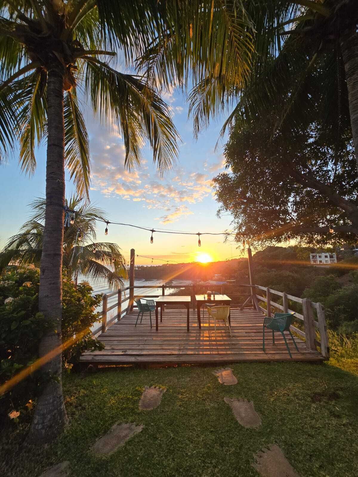 A wooden deck with tables and chairs, overlooking a body of water at sunset.