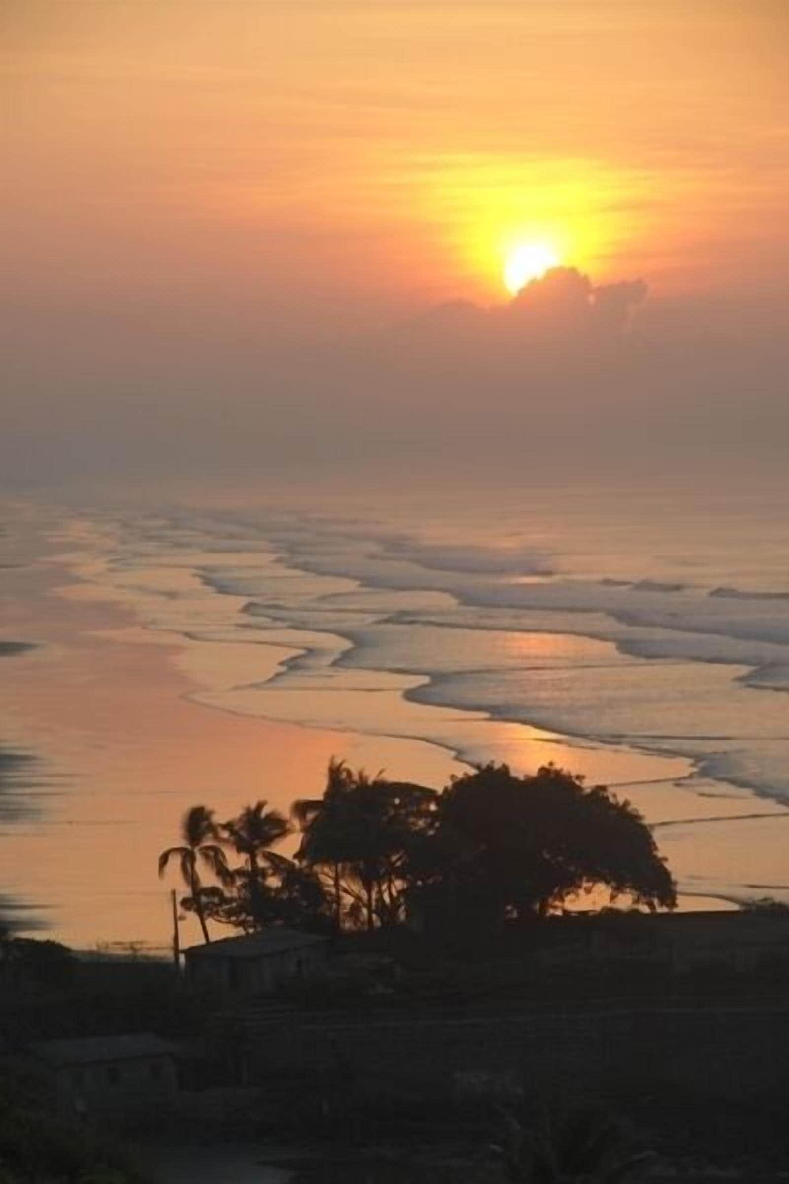 A sunset over a coastal area with waves and a silhouette of a house and palm trees.