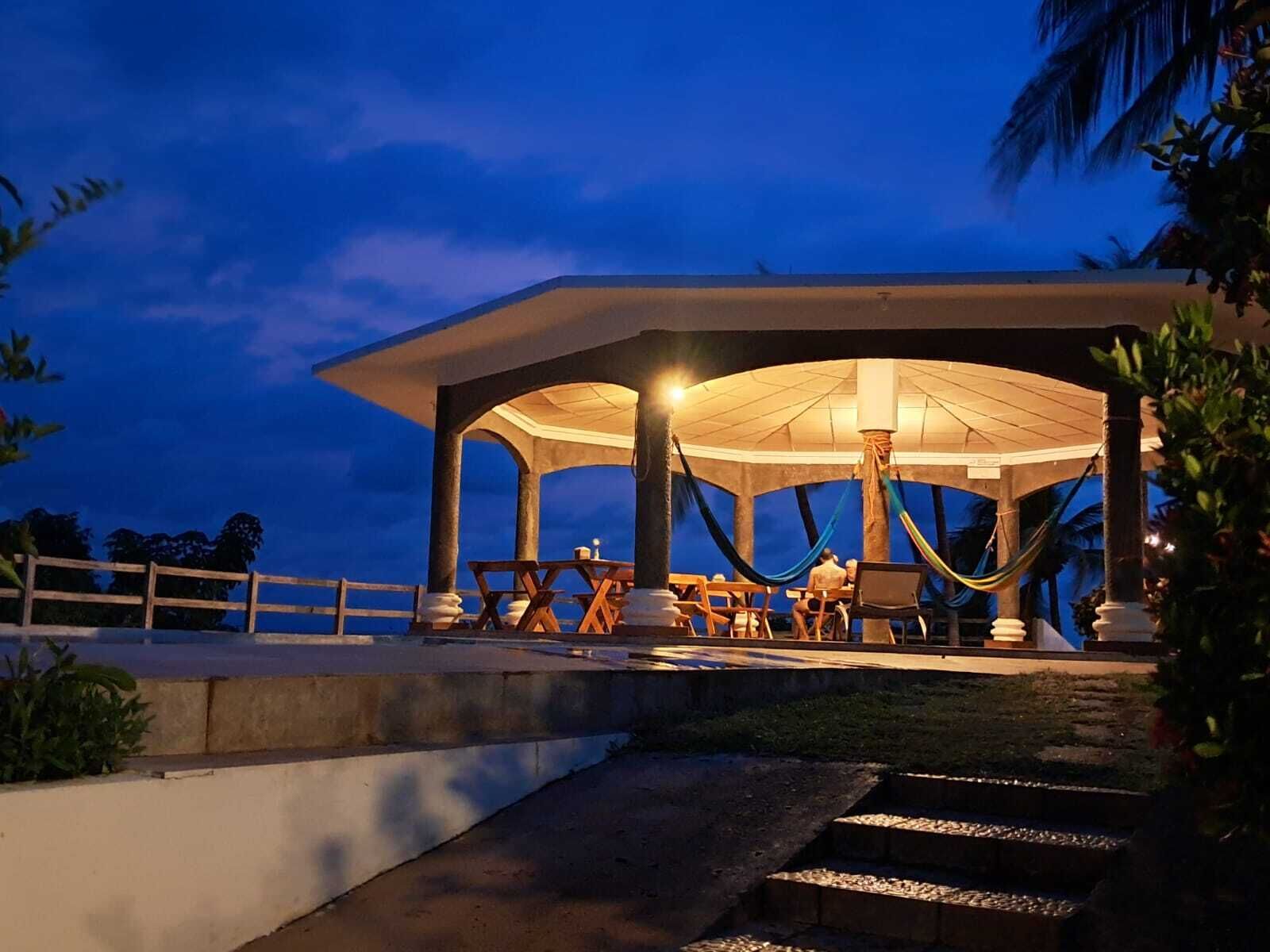 A covered outdoor seating area with tables and chairs, a hammock, and a view of the ocean at dusk.