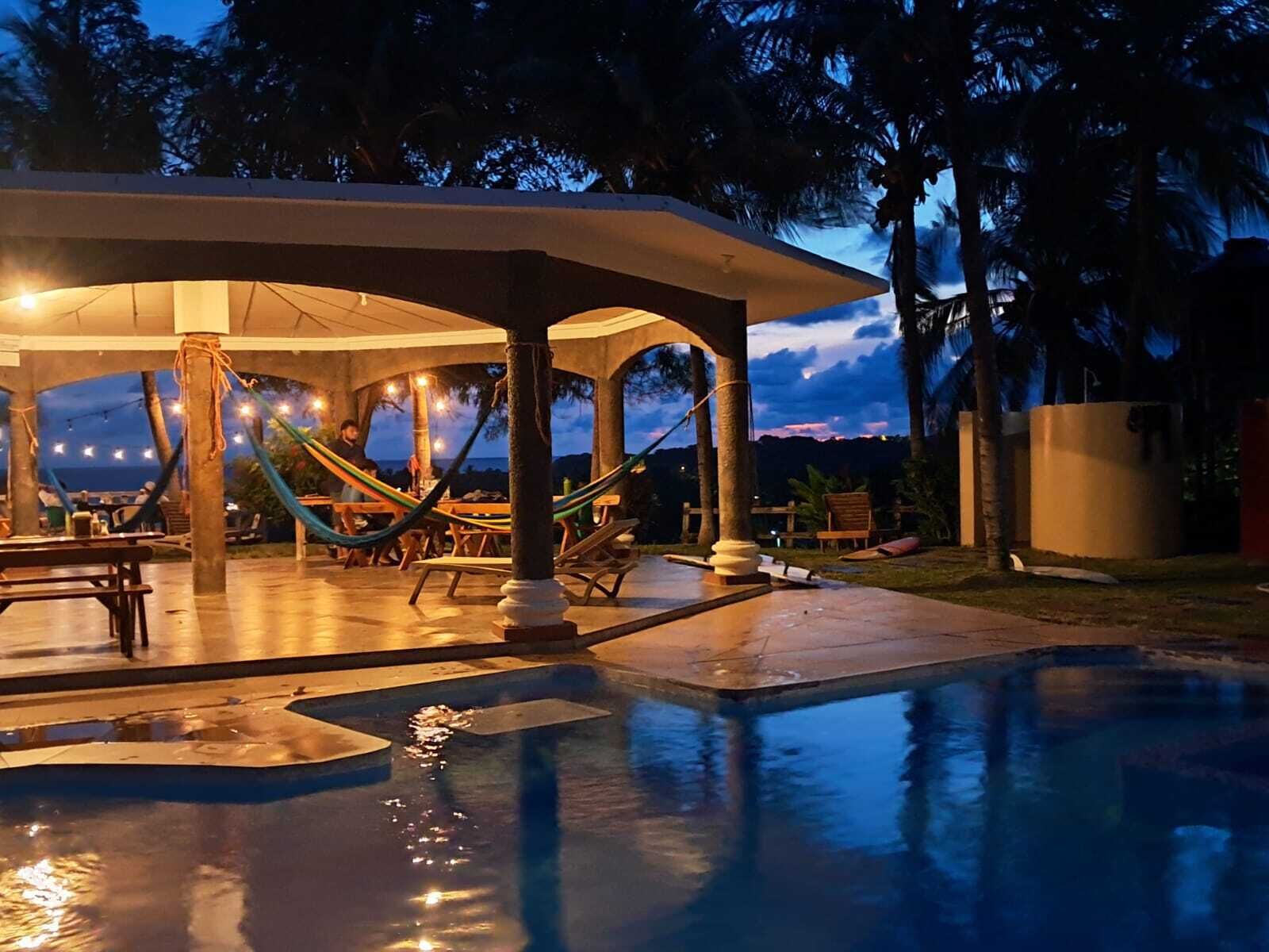 An outdoor pool area with a covered seating area, hammocks, and a view of the sky.