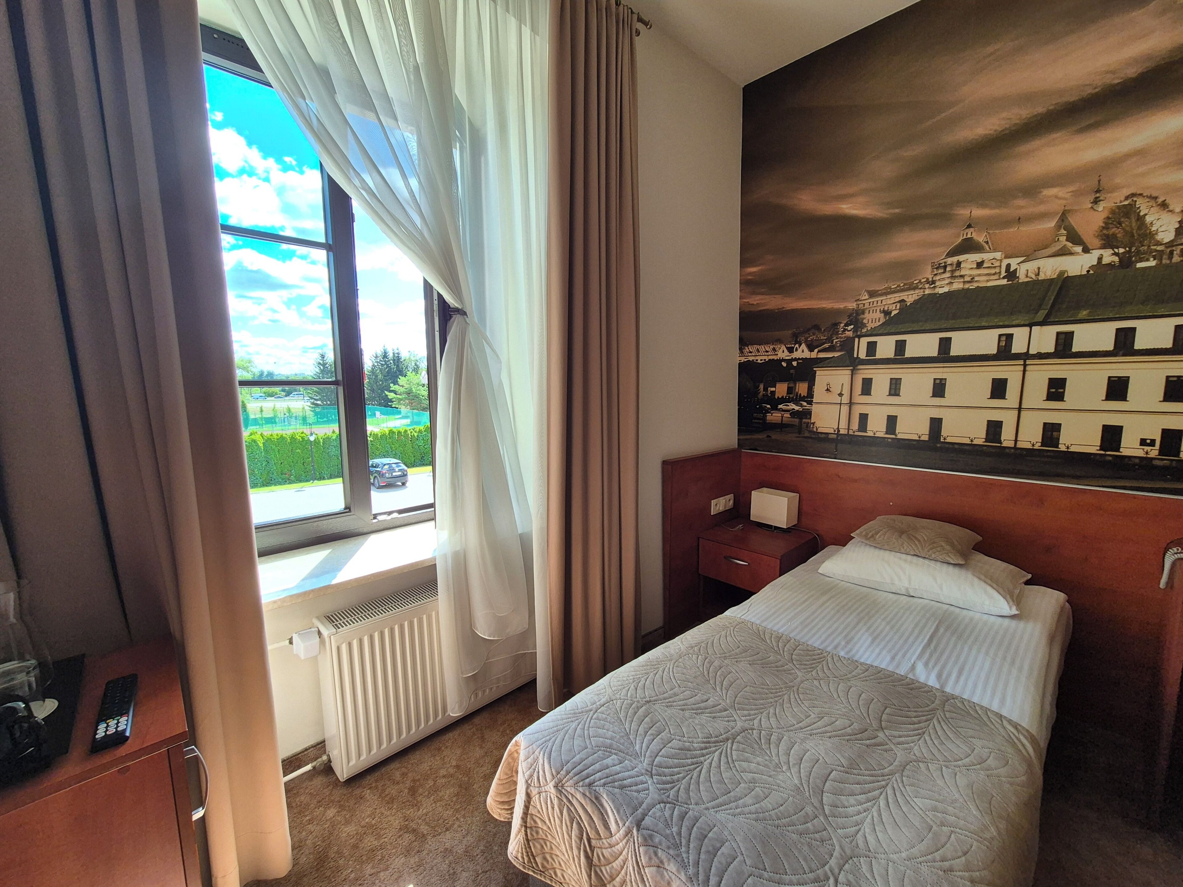 A hotel room with a bed, a nightstand, a radiator, a window with curtains, and a cityscape mural on the wall.