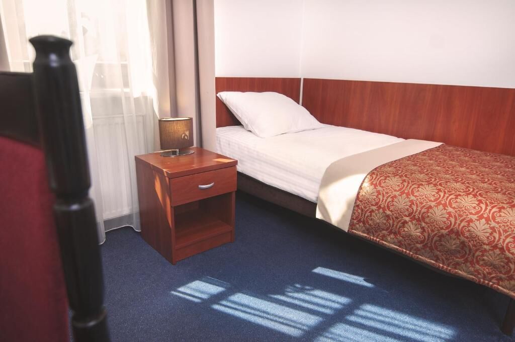 A hotel room with a single bed, a nightstand with a lamp, and a window with sheer curtains.