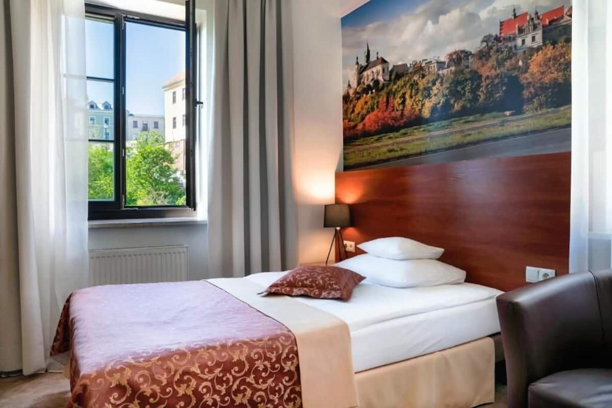 Una habitación de hotel con una cama, una ventana con cortinas, una lámpara y un cuadro de un castillo en la pared.