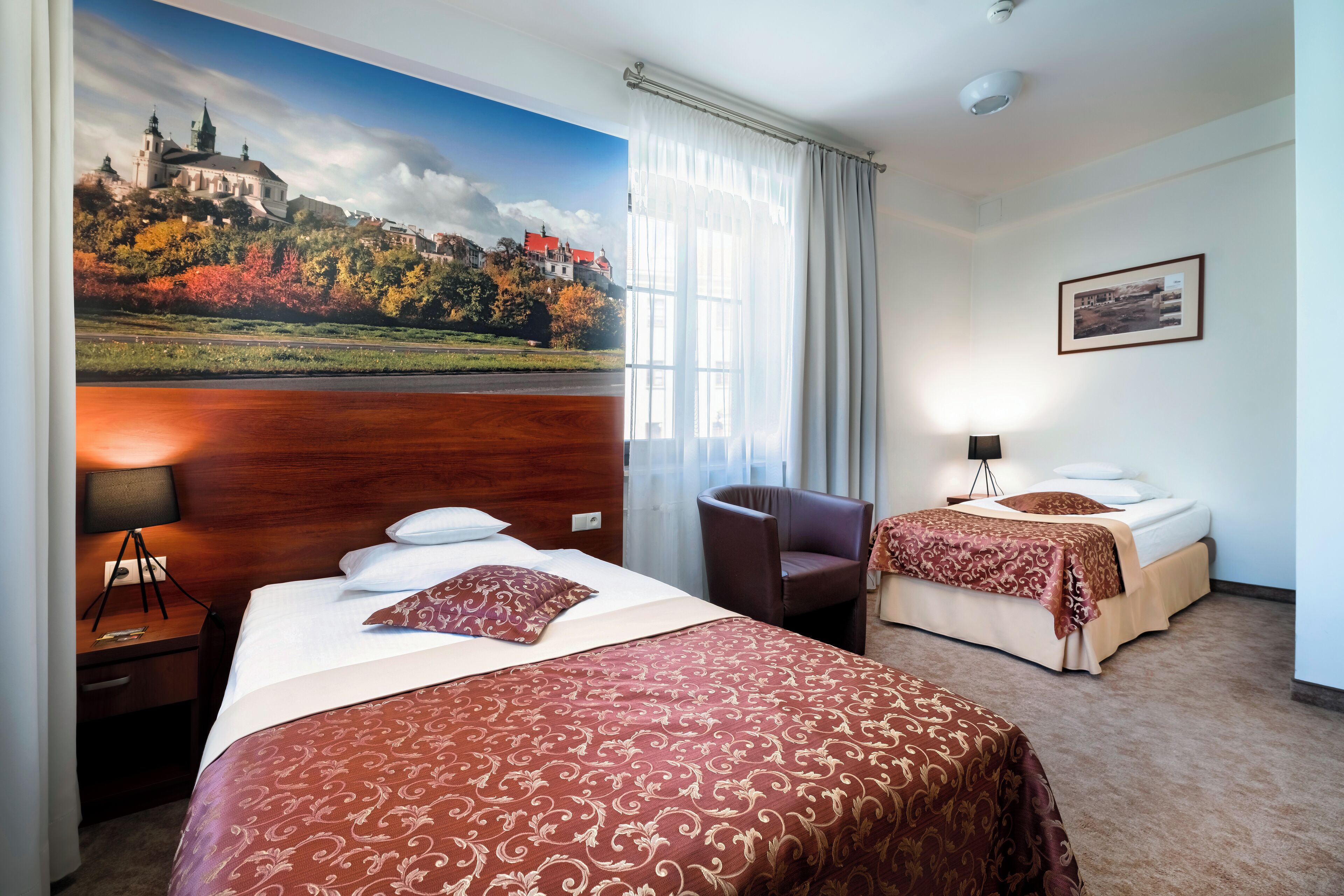 A hotel room with two beds, a large window with curtains, a desk with a lamp, and a painting of a castle on the wall.