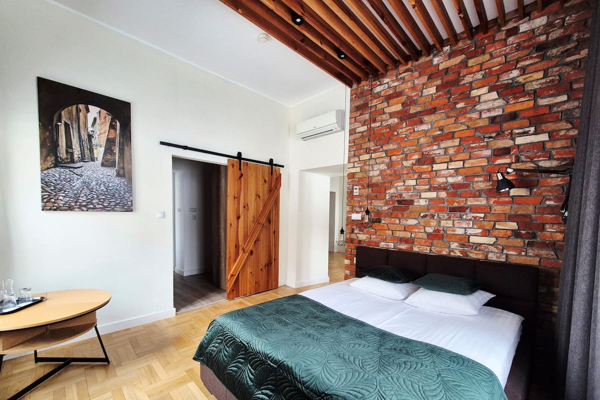 Un dormitorio con pared de ladrillo, una cama con colcha verde, una puerta de madera y un cuadro en la pared.