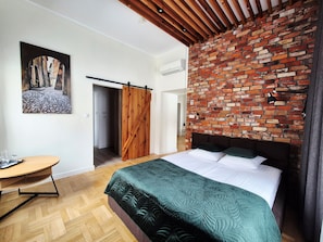 Une chambre avec un mur en briques, un lit recouvert d’une couette verte, une porte en bois et un tableau accroché au mur.