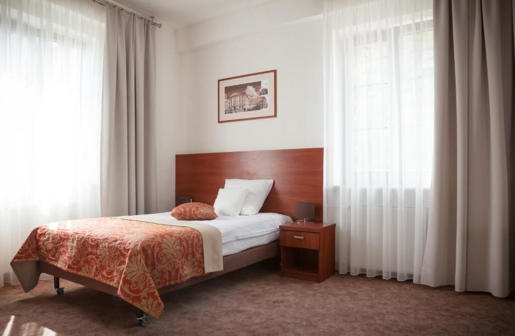 Una camera d'albergo con una testiera in legno, un letto con copripiumino a motivi rossi, un comodino con una lampada e tende leggere.