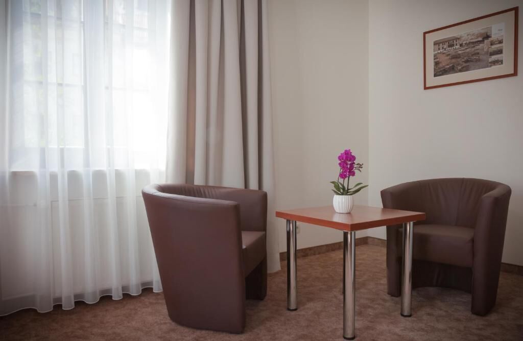 A room with two brown armchairs, a small wooden table with a white vase, and a window with sheer curtains.