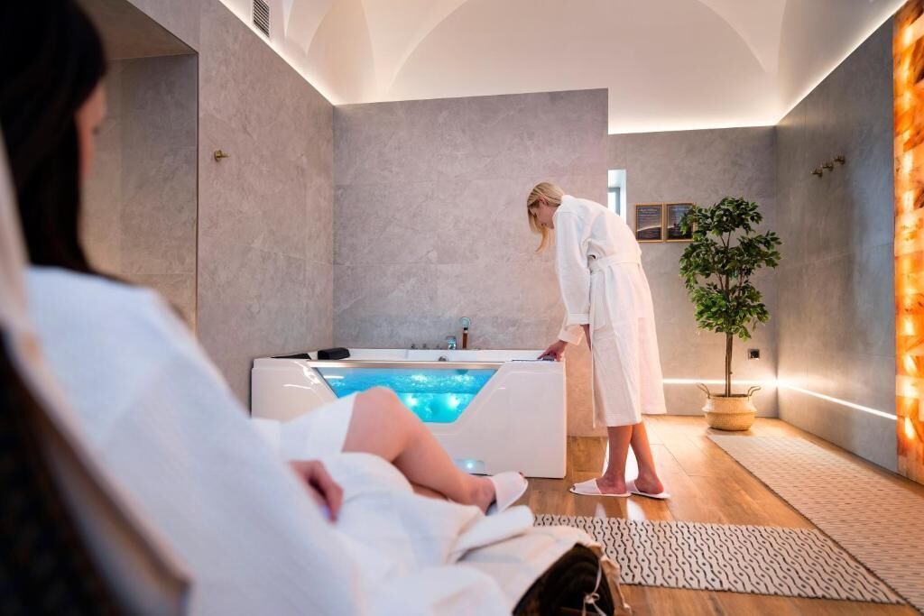 Um quarto de spa com uma banheira grande, uma planta em vaso e uma pessoa de roupão cuidando de outra pessoa deitada na banheira.