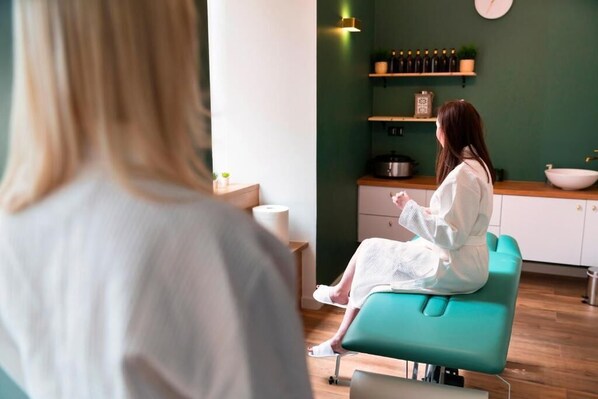 A person sitting on a teal chair in a room with a green wall, a wooden counter, and shelves with bottles. - Dom Na Podwalu Lublin (Lublin)