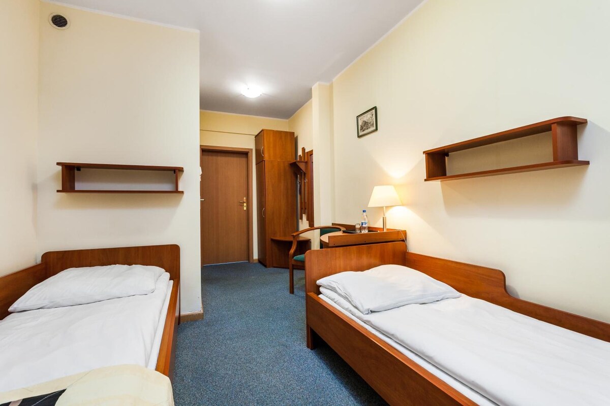 Habitació d'hotel amb dos llits individuals, una taula de treball amb una cadira, una tauleta de nit i una petita estantèlia.