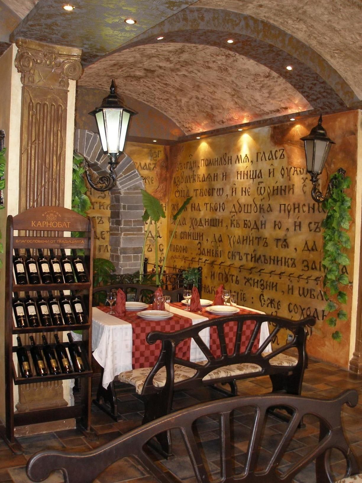 Interior de restaurante con una estantería de vinos, mesas, sillas y una pared con texto escrito a mano.