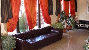 Una habitación con un sofá marrón oscuro, ventanas amplias con cortinas rojas y plantas en macetas.