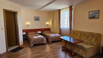 Habitación de hotel con dos camas individuales, un sofá, una mesa de madera, una puerta y dos luces fijadas a la pared.