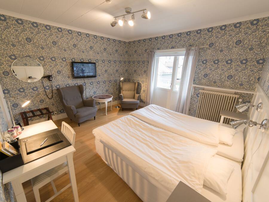 Un dormitorio con cama, un escritorio con silla, una mesita, una silla, televisor y ventana con cortinas.