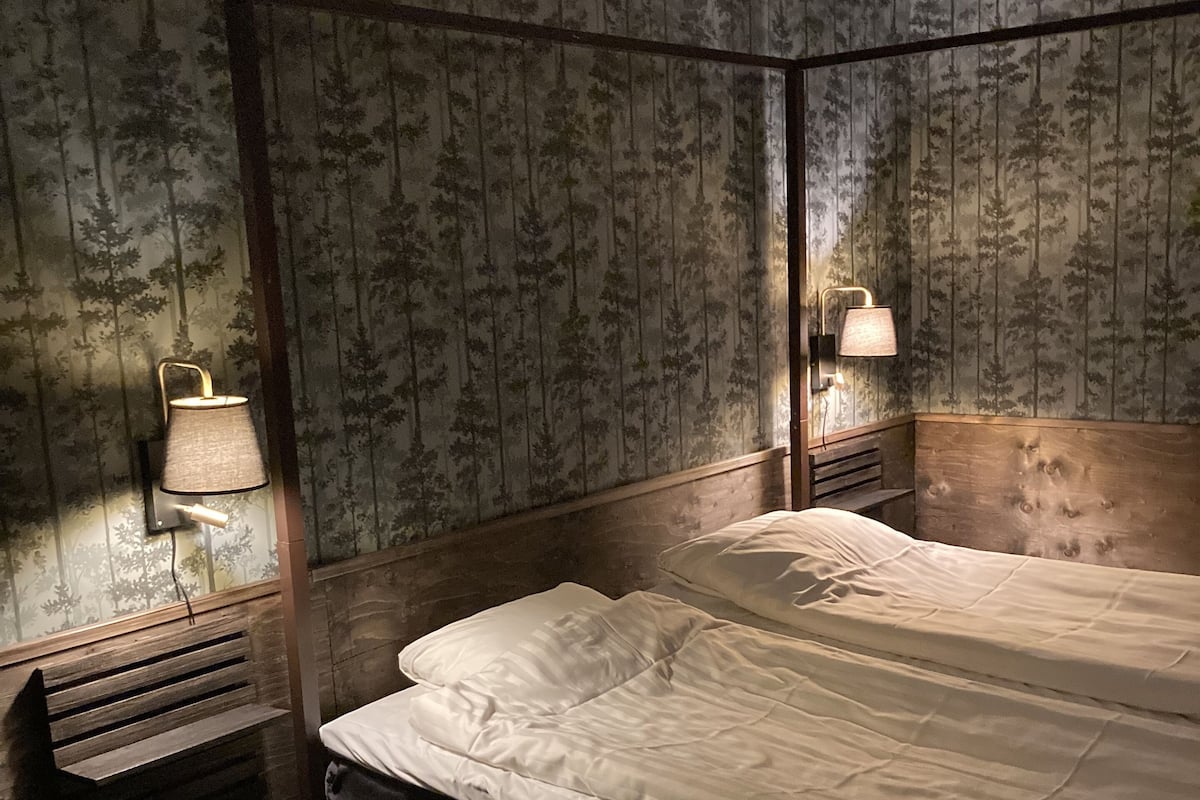 Ein Bett mit Baldachin, mit Holzrahmen und weißer Bettwäsche, flankiert von zwei Nachttischlampen und einer Bank. Der Raum zeichnet sich durch Tapeten mit vertikaler Musterung und Holzvertäfelung an den Wänden aus.