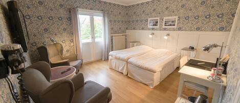 Habitación de hotel con cama, escritorio, silla, radiador y ventana con cortinas.