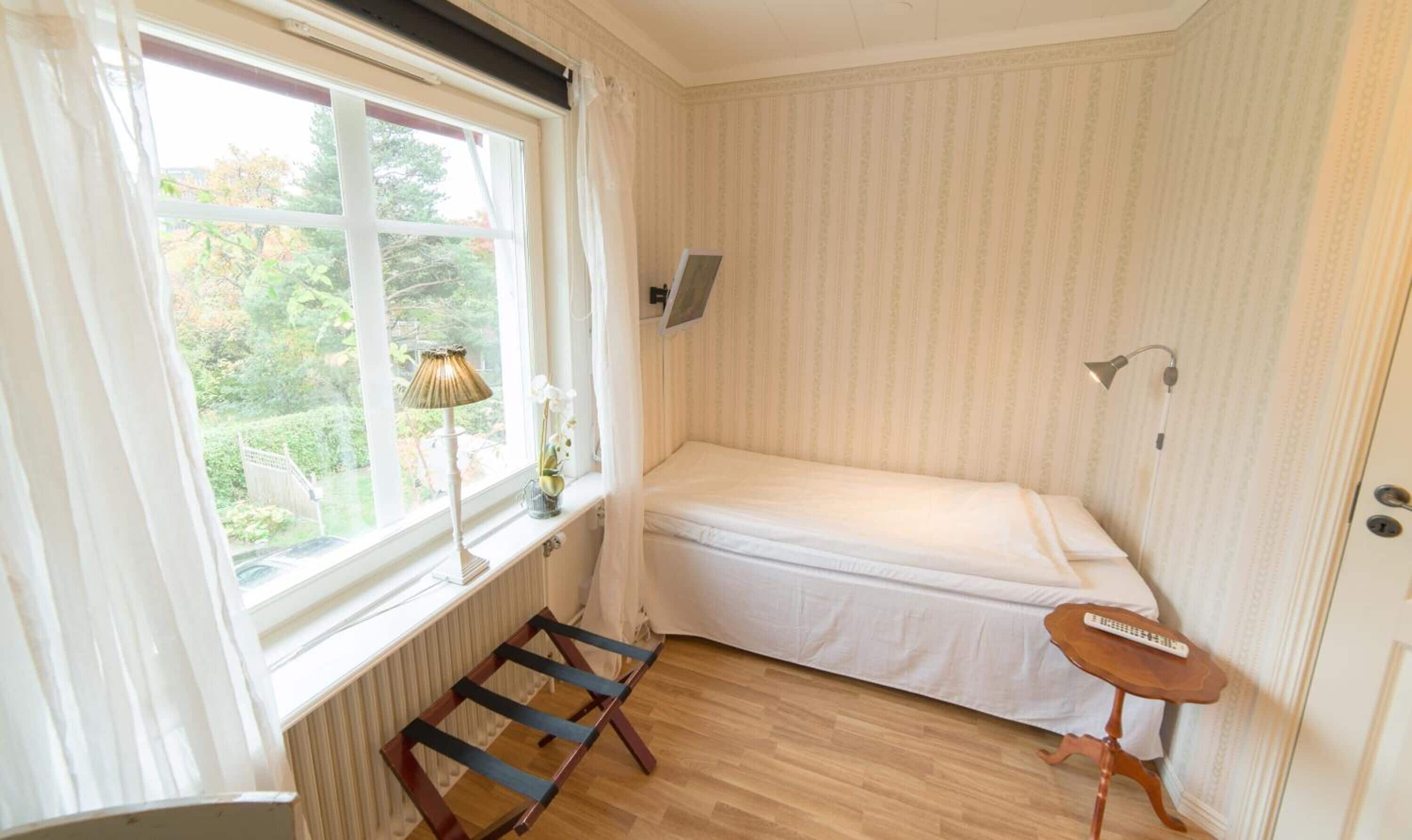 Une petite pièce, bien rangée, avec un lit simple, une fenêtre avec des rideaux, un radiateur, une petite table et une lampe.