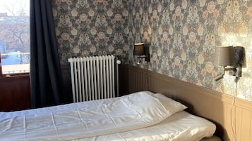 A bedroom with a bed, a radiator, a window with curtains, and a small table.