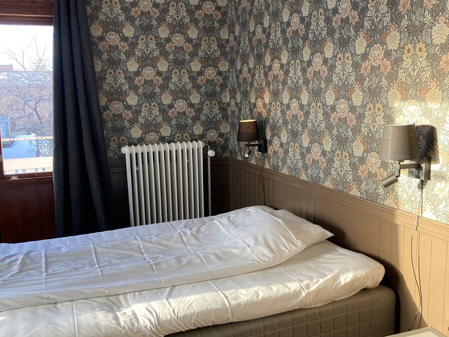 A bedroom with a bed, a radiator, a window with curtains, and a small table.