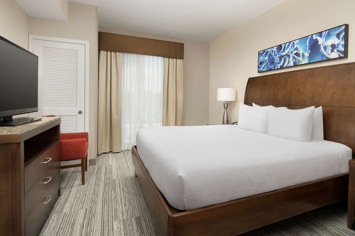 A hotel room with a bed, a television, a desk, a chair, a lamp, and a painting on the wall.