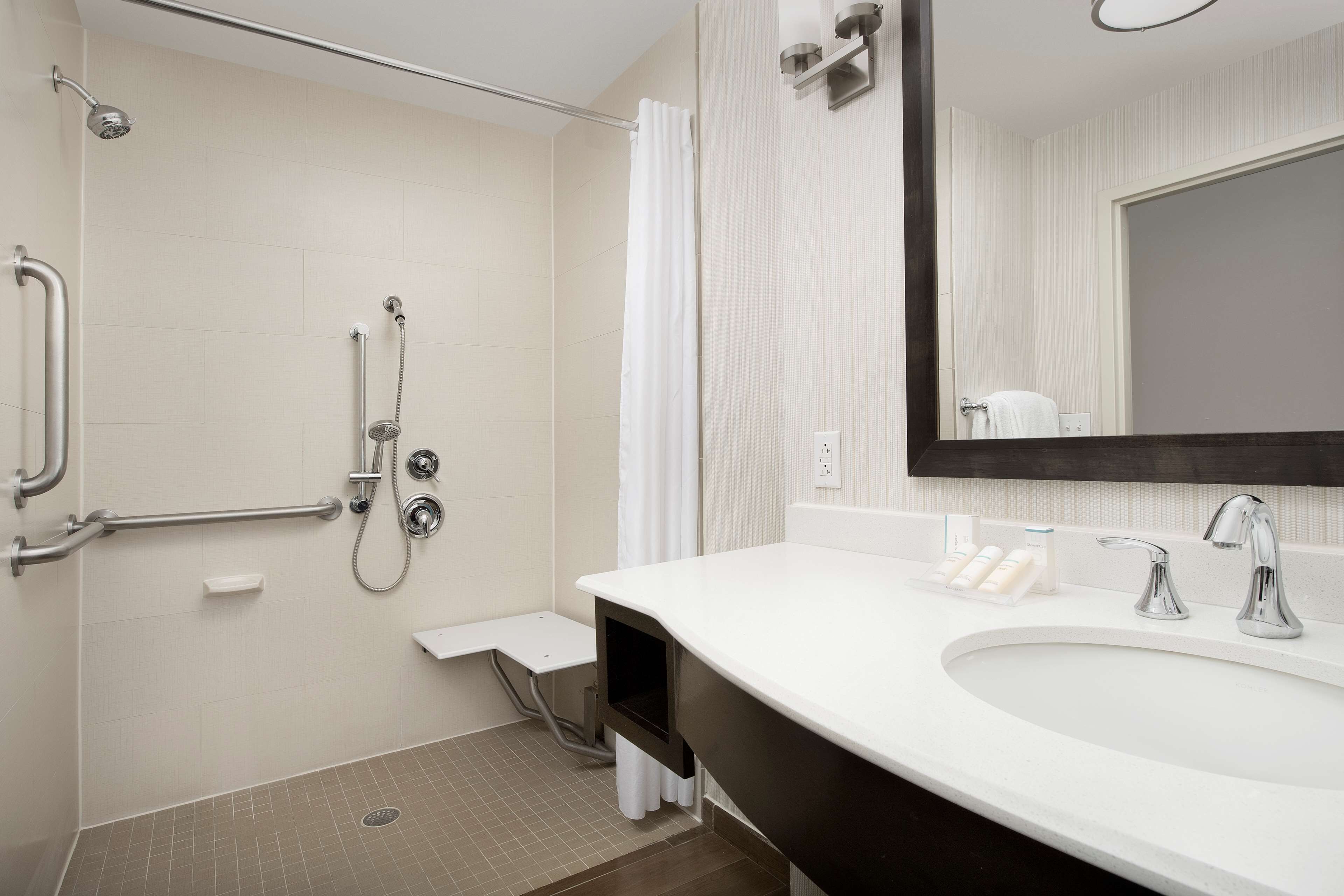 Un bagno moderno con un lavandino bianco, uno specchio grande e una doccia dotata di maniglie di sostegno.