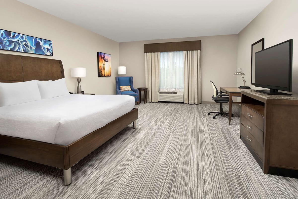 Ein Hotelzimmer mit einem großen Bett, einem Schreibtisch mit Computer, einem Fernseher, einem Sessel und Bildern an den Wänden.