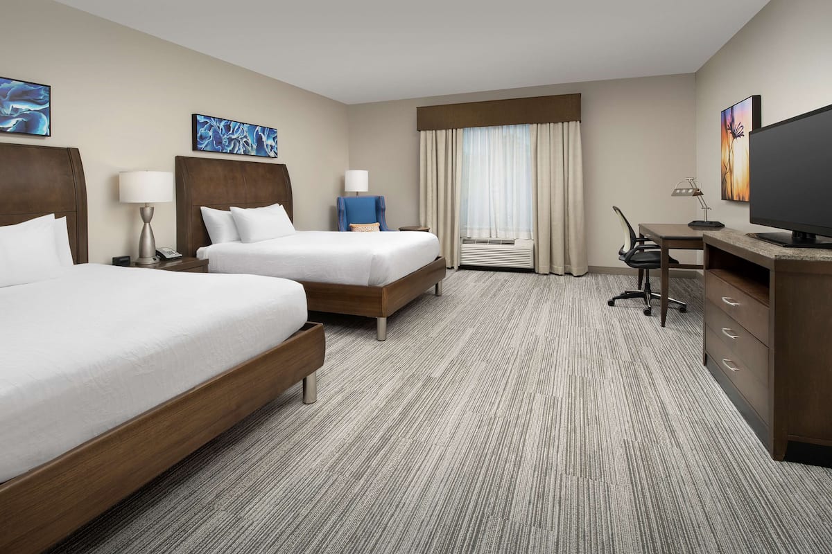 A hotel room with two beds, a desk, a chair, a television, and artwork on the walls.