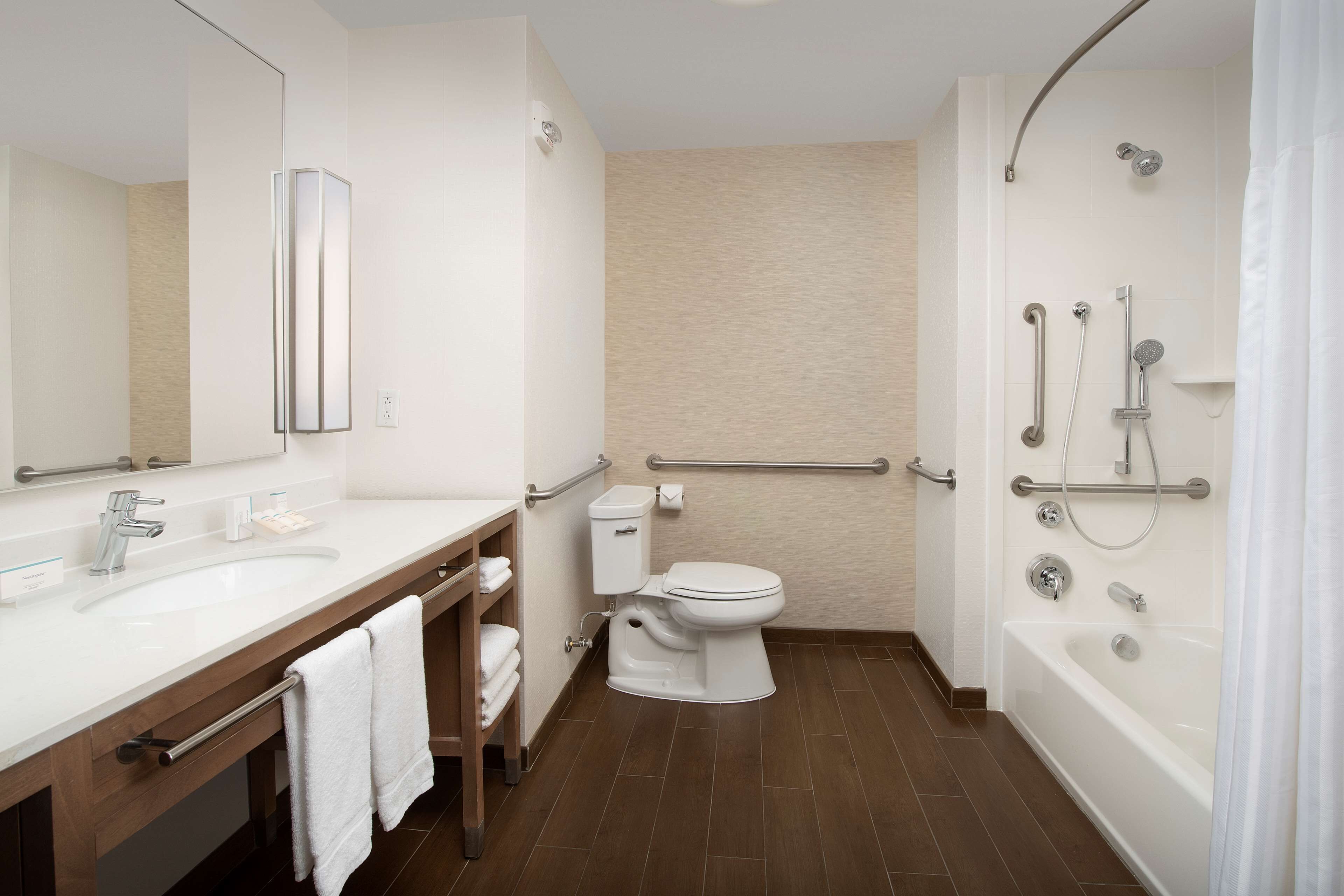 A modern bathroom with a white sink, toilet, and bathtub. It includes a large mirror, a towel rack, and a shower with grab bars.