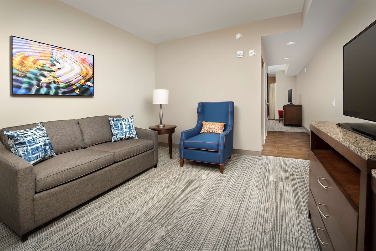 A modern hotel room with a gray sofa, a blue armchair, a flat-screen TV, and a large abstract painting on the wall.