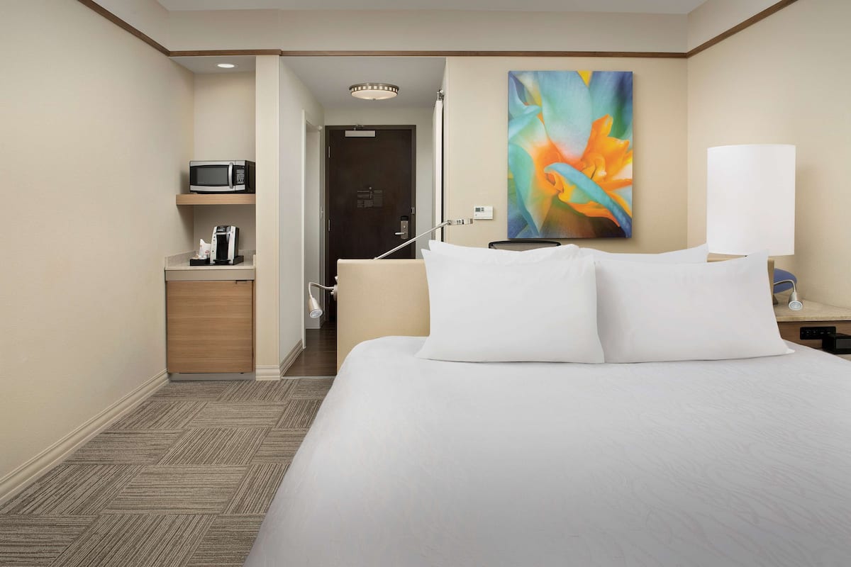 A hotel room with a bed, a microwave, a coffee maker, a painting, and a lamp.