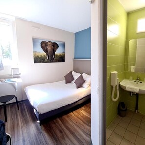 Une chambre à coucher avec un lit, un bureau, une chaise, un tableau représentant un éléphant, et une salle de bain carrelée de carreaux verts.