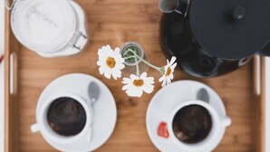 Una bandeja de madera con dos tazas blancas de café, una cafetera negra, una jarrita de leche blanca y un tarro pequeño con flores blancas.