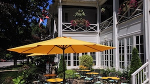 An outdoor patio with yellow umbrellas, wicker chairs, and tables, surrounded by greenery and a building with large windows.