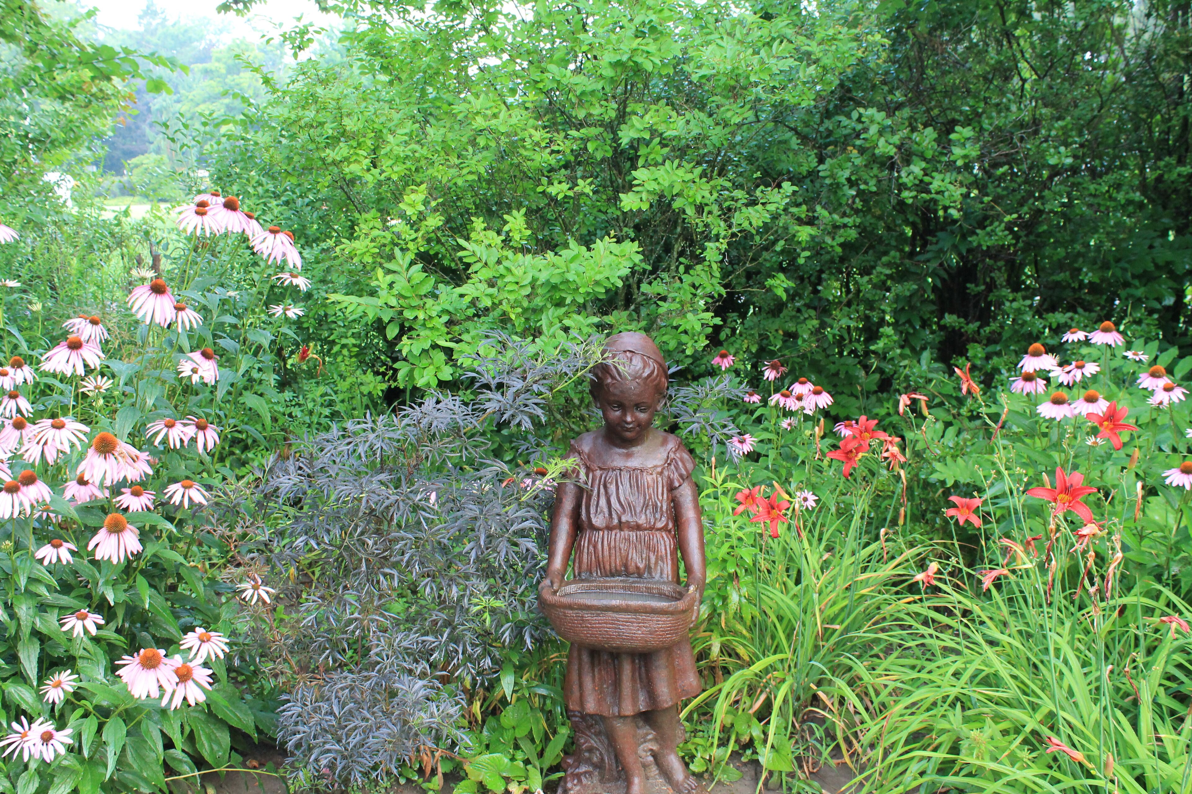 A garden statue of a girl holding a basket amidst various flowers and greenery.