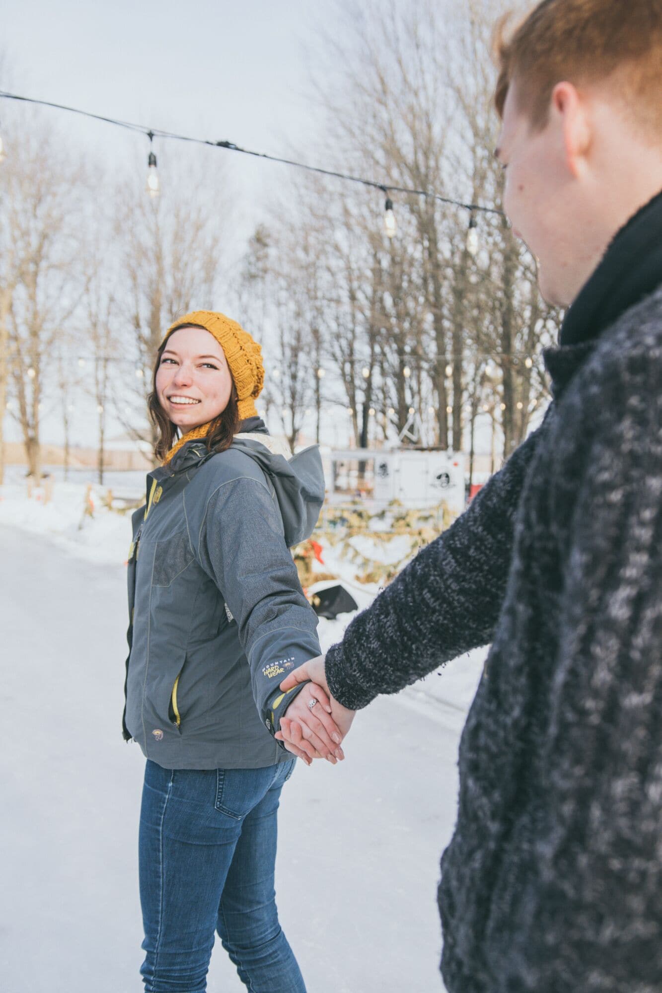 Two people holding hands in a snowy outdoor setting with bare trees and string lights in the background.