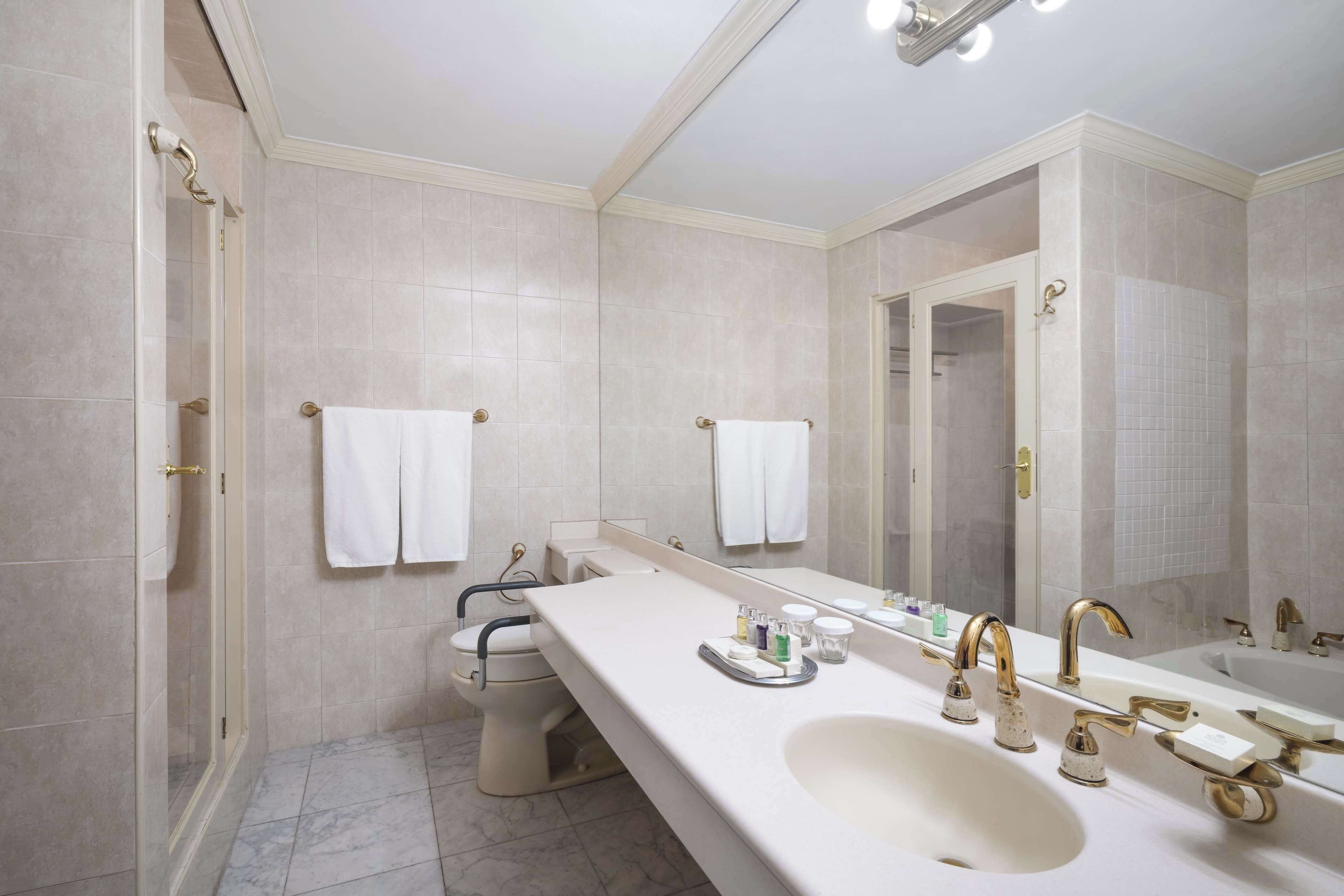 A modern bathroom with a large sink, two gold faucets, a mirror, and a toilet.