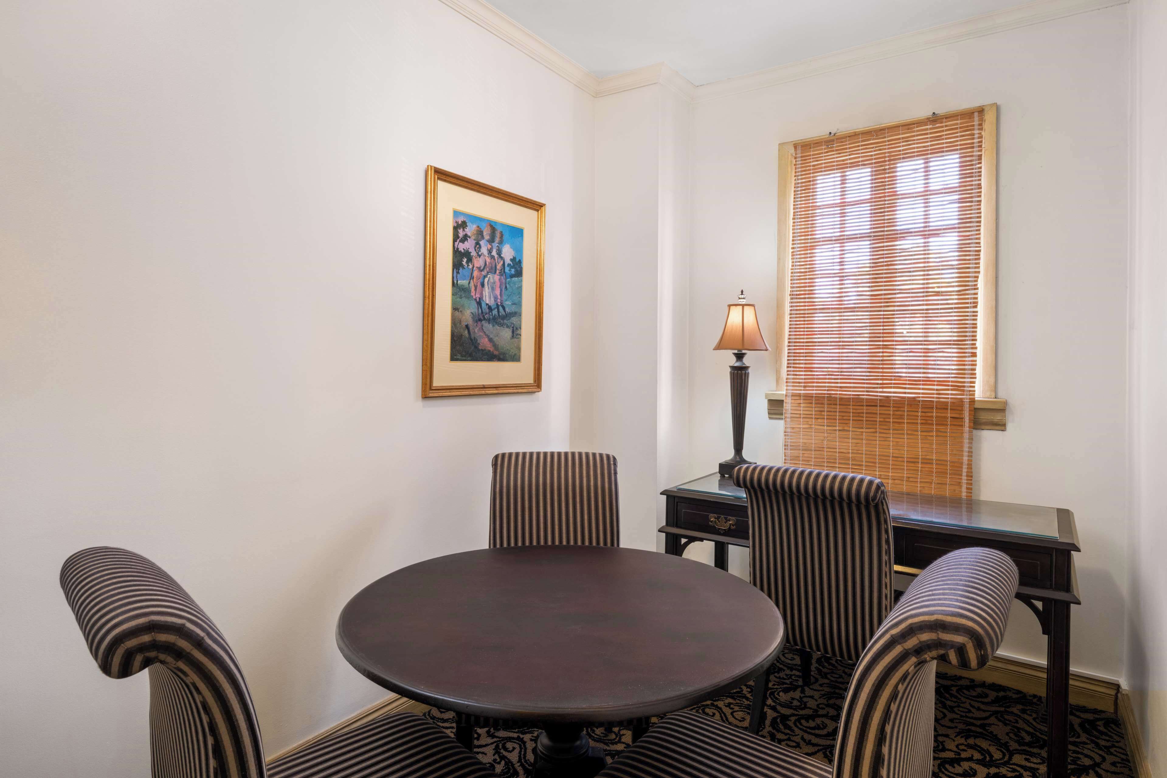 A dining area with a round table, four striped chairs, a lamp, a framed picture on the wall, and a window with blinds.