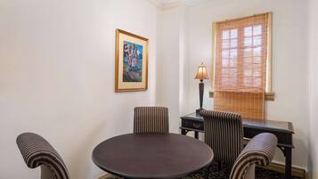 Un coin repas avec une table ronde, quatre chaises à rayures, une lampe, un tableau encadré au mur et une fenêtre avec des stores.
